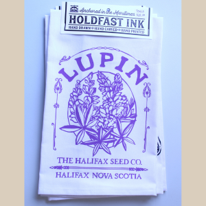 A photo of a white tea towel with a purple design of lupins with purple text reading Lupin - The Halifax Seed Co. - Halifax Nova Scotia