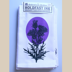 a photo of a white tea towel with a screen printed image of a black scottish thistle with a purple background